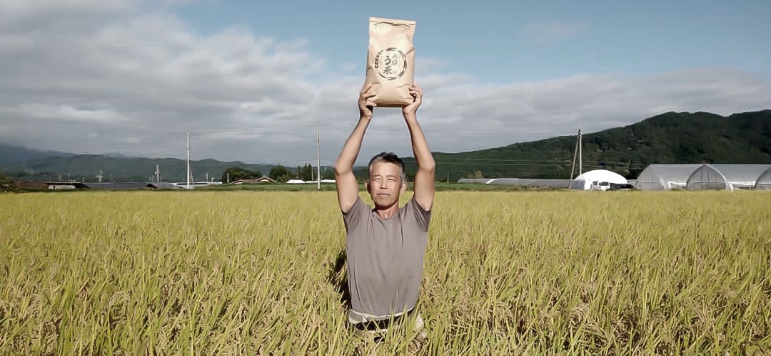 代表が一面黄金色の田んぼでコメ袋を両手で持ち上げる風景